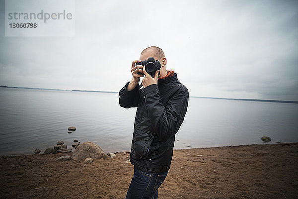 Mann fotografiert mit Kamera  während er am Seeufer vor bewölktem Himmel steht