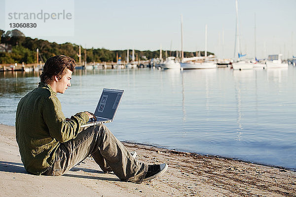 Mann benutzt Laptop-Computer  während er am Strand vor klarem Himmel sitzt