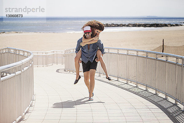 Glücklicher Mann nimmt Frau huckepack  während er am Strand auf der Strandpromenade spazieren geht
