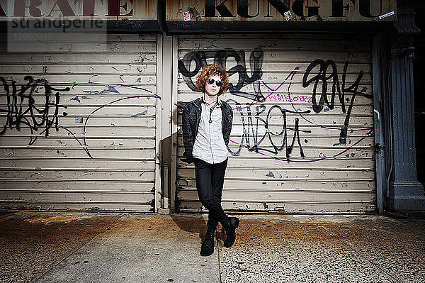 Porträt eines jungen Mannes  der auf einem Fußweg vor Graffiti-Fensterläden steht