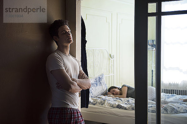 Nachdenklicher Mann lehnt an der Wand  während seine Freundin im Schlafzimmer schläft
