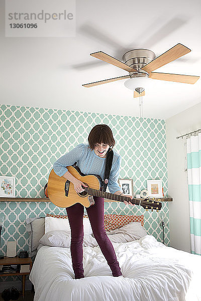 Glückliche Frau spielt Gitarre  während sie zu Hause auf dem Bett steht