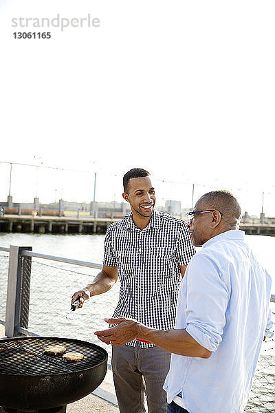Lächelnder Mann bereitet Essen zu  während er mit seinem Vater am East River vor klarem Himmel steht