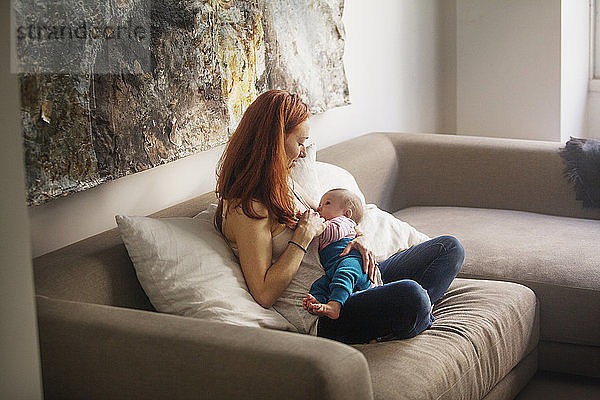 Mutter füttert kleines Mädchen  während sie zu Hause auf dem Sofa sitzt