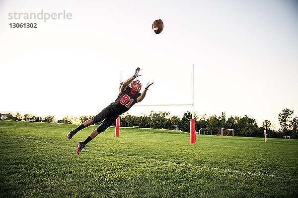 Junger Mann taucht  um American Football auf dem Spielfeld gegen den Himmel zu fangen
