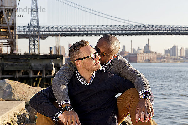 Mann küsst seinen Freund  während er gegen die Williamsburg Bridge sitzt