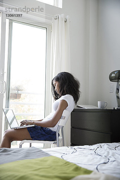 Seitenansicht einer Frau  die einen Laptop benutzt  während sie zu Hause auf einem Stuhl am Fenster sitzt