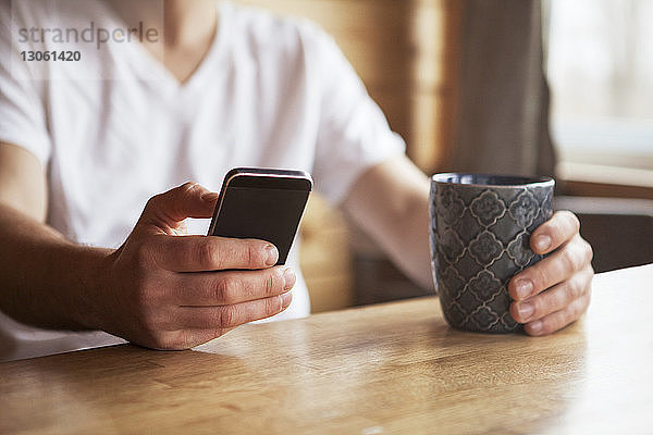 Mittelsektion eines Mannes  der ein Mobiltelefon benutzt  während er im Café eine Kaffeetasse hält