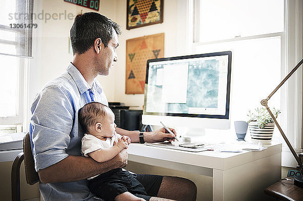 Mann trägt einen kleinen Jungen  während er zu Hause am Desktop-Computer arbeitet