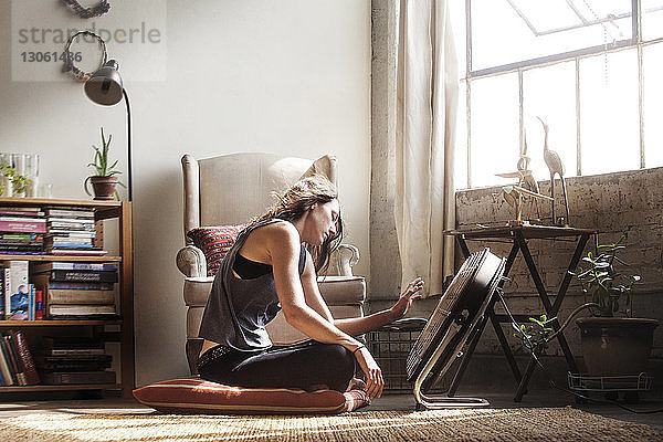 Seitenansicht einer Frau  die zu Hause vor einem elektrischen Ventilator sitzt