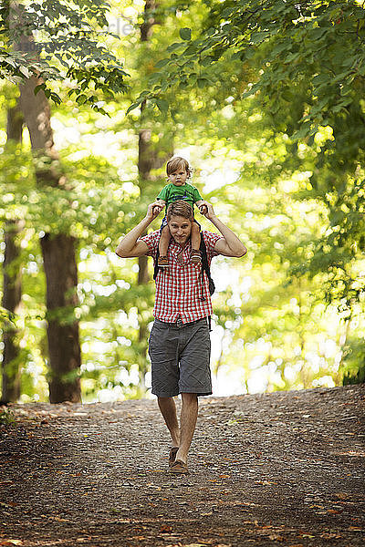 Porträt eines Vaters  der seinen Sohn auf den Schultern trägt  während er im Wald auf einem Feldweg geht