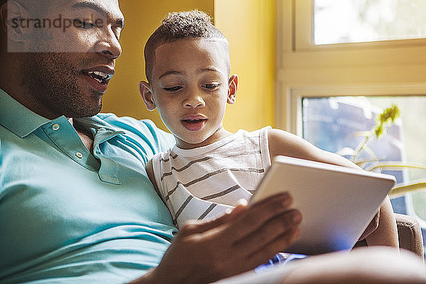 Mann zeigt seinem Sohn einen Tablet-Computer  während er zu Hause auf dem Sofa sitzt