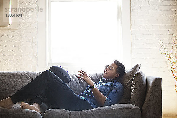 Mann hält Rugbyball  während er zu Hause auf dem Sofa liegt