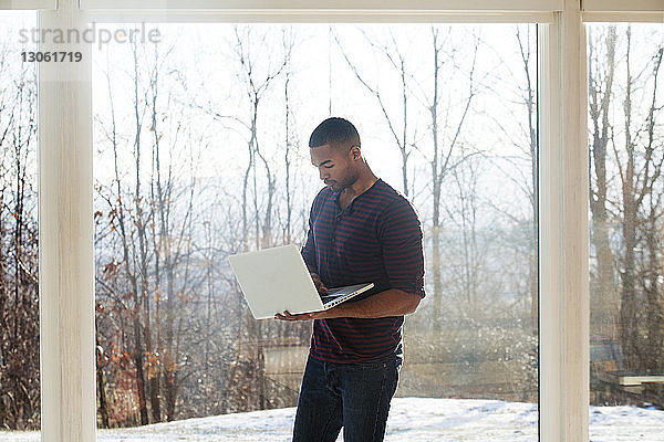 Mann benutzt Laptop-Computer  während er zu Hause gegen ein Glasfenster steht