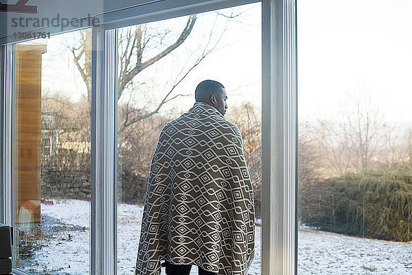 Rückansicht eines mit einer Decke bedeckten Mannes  der zu Hause am Fenster steht
