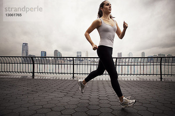 Seitenansicht einer Sportlerin  die auf einem Gehweg am Fluss und bei bewölktem Himmel joggt