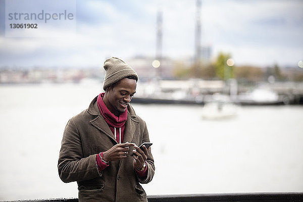 Lächelnder Mann benutzt Mobiltelefon  während er an der Reling gegen das Meer steht