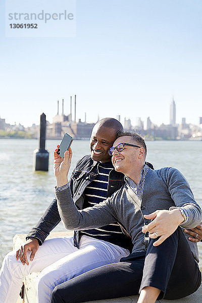 Glückliche Freunde nehmen Selfie am Fluss gegen den klaren Himmel an einem sonnigen Tag