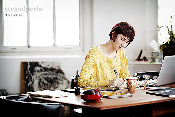 Geschäftsfrau schreibt in Buch  während sie am Laptop am Schreibtisch im Kreativbüro arbeitet