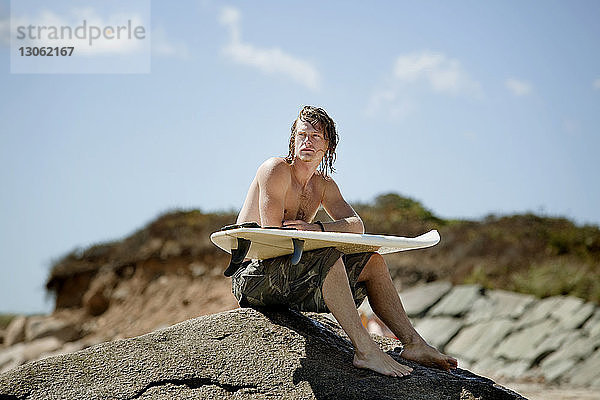 Mann mit Surfbrett schaut weg  während er auf einem Felsen sitzt