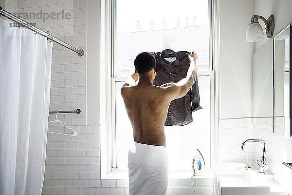 Rückansicht eines Mannes  der im Badezimmer ein gebügeltes Hemd am Fenster hält