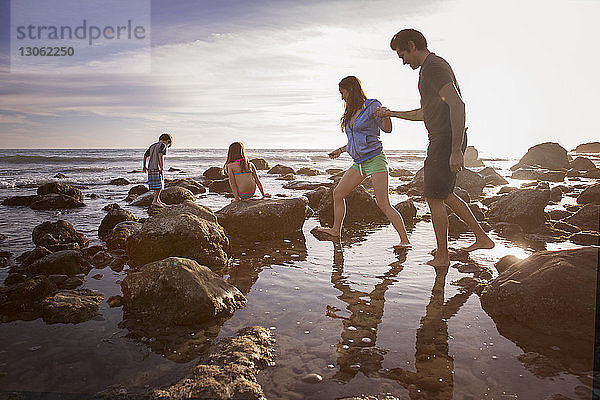 Paar hält sich an den Händen  während Kinder am Strand am Felsen spielen