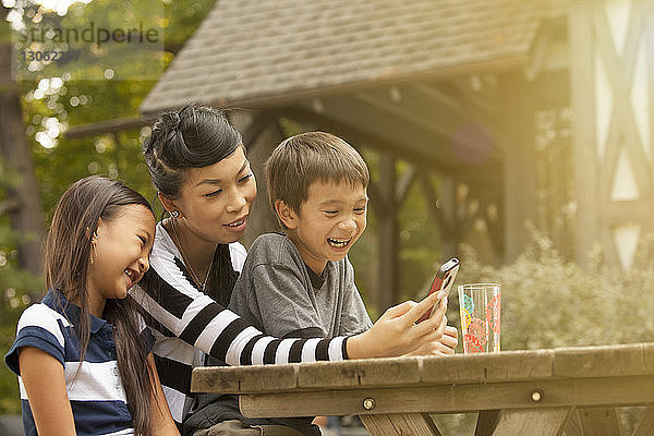 Glückliche Mutter und Kinder telefonieren  während sie im Garten am Tisch sitzen