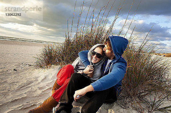 Mann küsst Freundin  während er am Strand vor bewölktem Himmel sitzt
