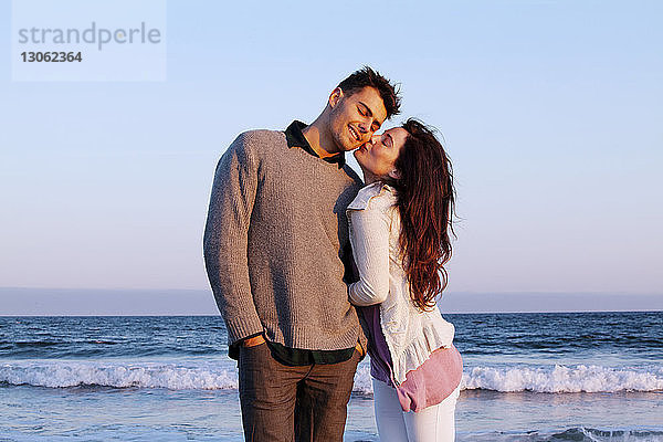 Glückliche Frau küsst Mann  während sie am Strand vor klarem Himmel steht