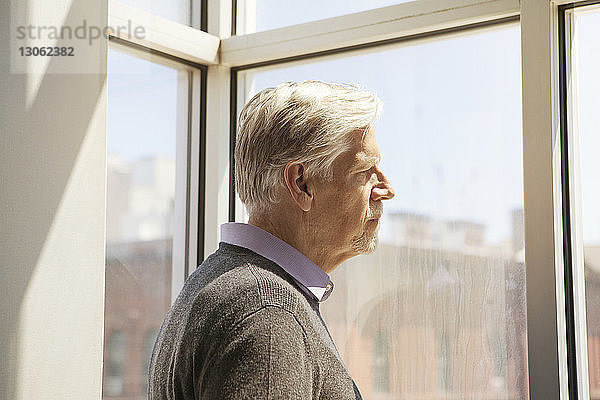 Seitenansicht eines älteren Mannes  der zu Hause durch ein Fenster schaut