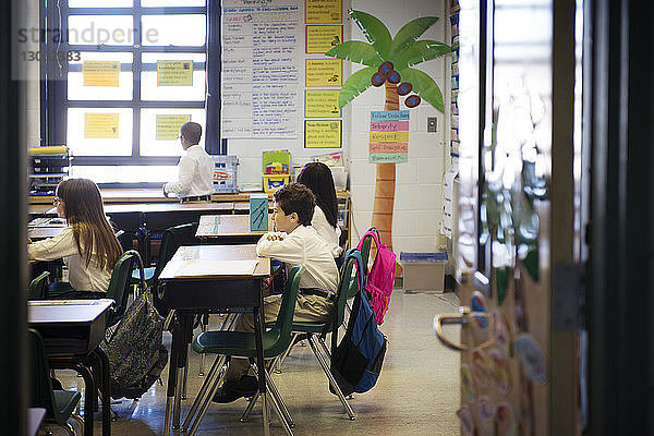 Kinder  die an Schreibtischen im Klassenzimmer sitzen und durch eine Tür gesehen werden