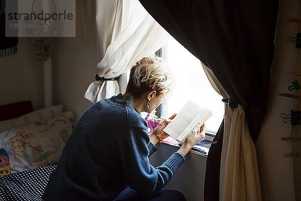 Hochwinkelansicht einer Frau  die ein Buch liest  während sie zu Hause am Fenster sitzt