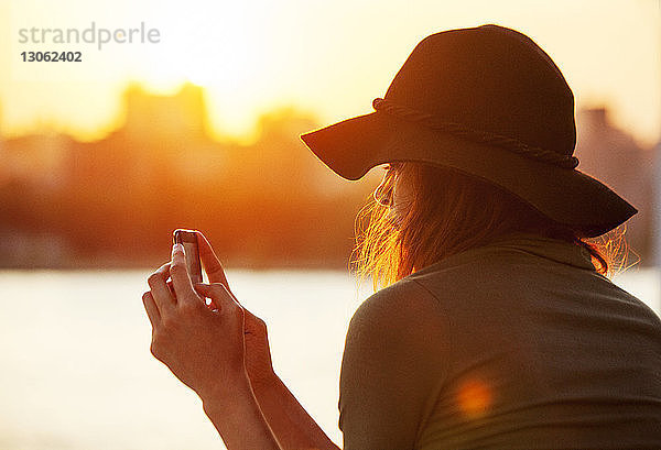 Rückansicht einer Frau  die während des Sonnenuntergangs ein Mobiltelefon benutzt