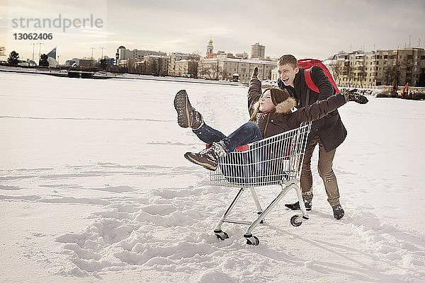 Glücklicher Mann schiebt seine Freundin  die im Einkaufswagen auf einem schneebedeckten Feld sitzt