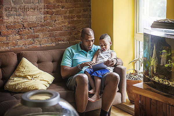 Vater zeigt seinem Sohn einen Tablet-Computer  während er zu Hause auf dem Sofa sitzt