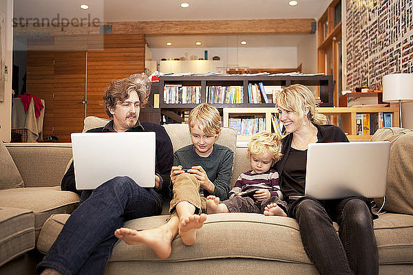 Geschwister benutzen Mobiltelefone  während sie mit den Eltern zu Hause auf dem Sofa sitzen