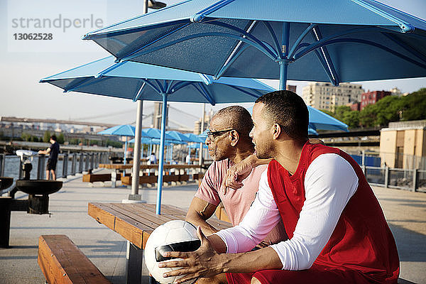 Vater sitzt mit Mann  der einen Fussball unter dem Sonnenschirm hält