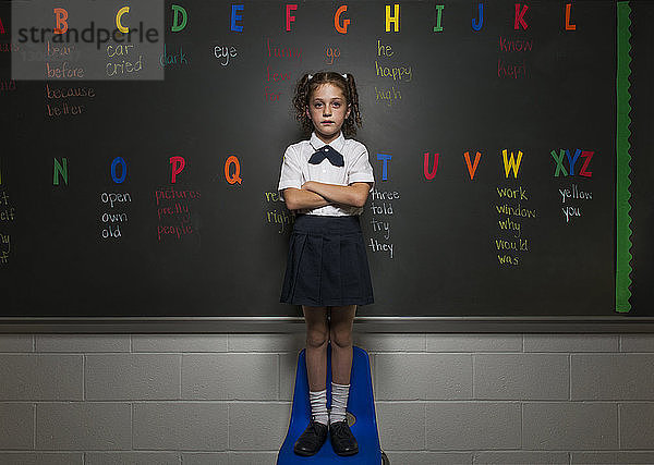 Porträt eines selbstbewussten Schulmädchens  das auf einem Stuhl gegen eine Tafel steht