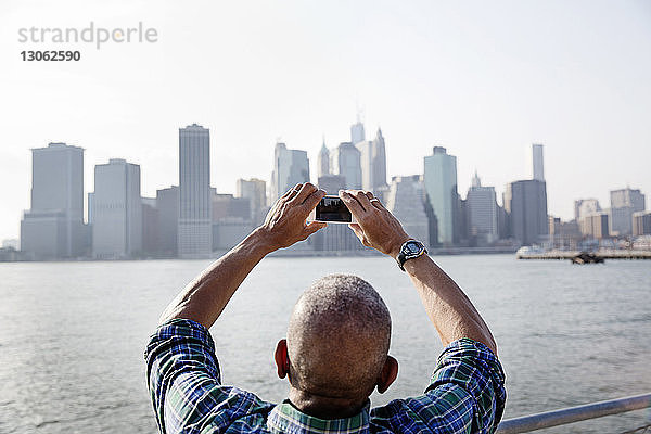 Rückansicht eines älteren Mannes  der die Stadt mit einem Smartphone am East River fotografiert