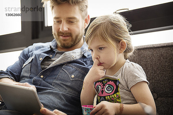 Mädchen sieht Vater am Tablet-Computer an  während sie zu Hause auf dem Sofa sitzt