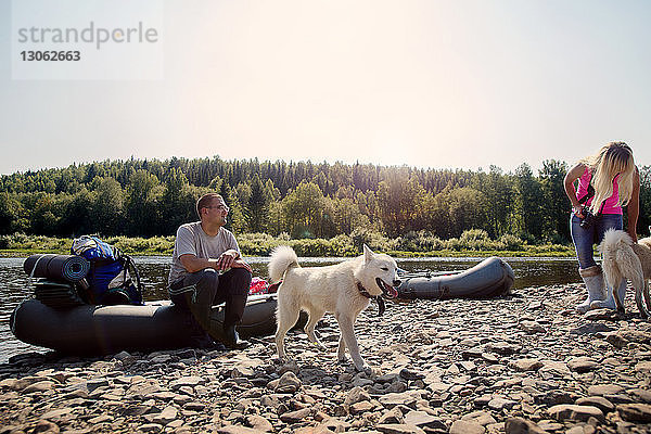 Freunde mit Hunden mit aufblasbaren Flößen am Flussufer bei klarem Himmel