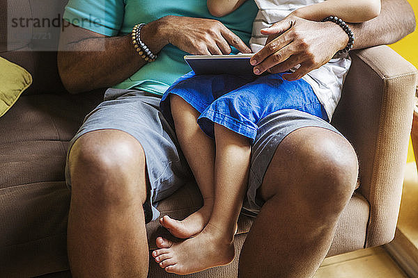 Mittelteil eines Vaters mit Sohn  der einen Tablet-Computer benutzt  während er zu Hause auf dem Sofa sitzt