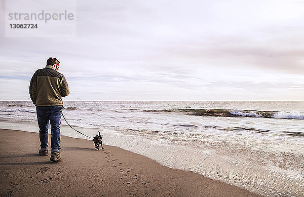 Rückansicht eines Mannes mit Hund beim Spaziergang am Ufer vor bewölktem Himmel