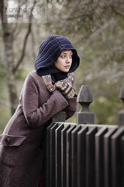 Frau in warmer Kleidung schaut weg  während sie sich an den Zaun lehnt