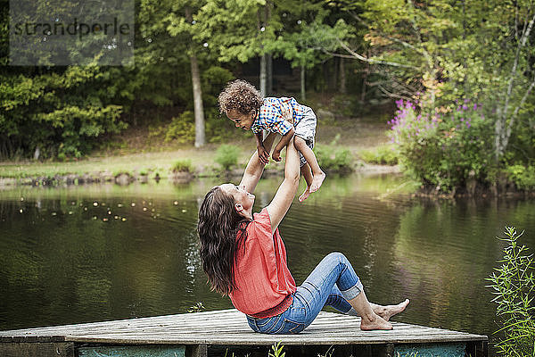 Glückliche Frau hebt Sohn hoch  während sie auf dem Steg am See sitzt