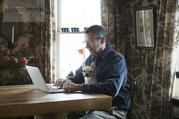 Mann benutzt Laptop-Computer  während er zu Hause mit Hund am Tisch sitzt