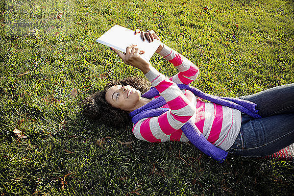 Hochwinkelansicht einer glücklichen Frau  die einen Tablet-Computer benutzt  während sie im Park liegt