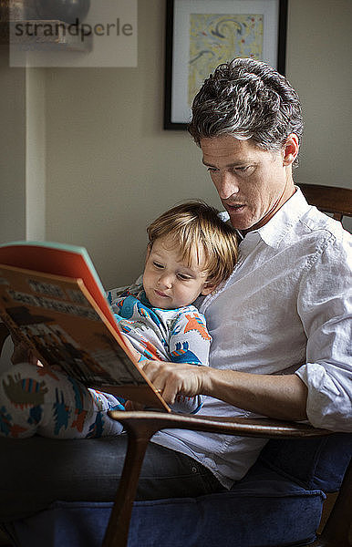 Vater liest dem Sohn ein Bilderbuch vor  während er zu Hause auf einem Stuhl sitzt