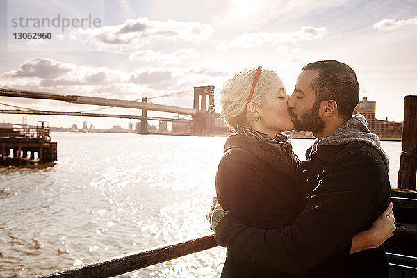 Ein Paar küsst sich  während es an einem sonnigen Tag an der Brooklyn Bridge über den East River steht