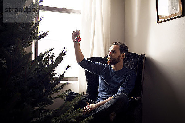 Mann betrachtet Weihnachtsschmuck  während er zu Hause auf einem Sessel am Weihnachtsbaum sitzt